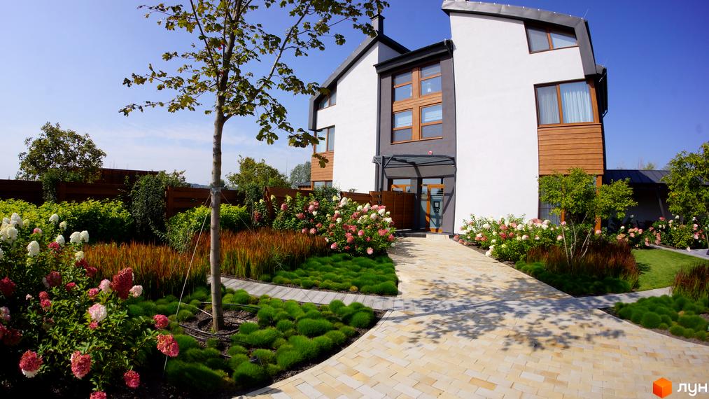 Зображення фасаду будинку з білими стінами та дерев'яними вставками. На передньому плані доглянута прибудинкова територія з рослинами та доріжкою з бруківки.