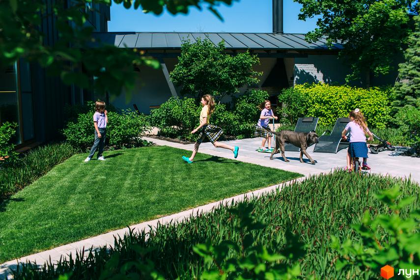 Будинок з темним дахом та великим двором. Діти граються на території: бігають, катаються на самокатах та велосипедах.