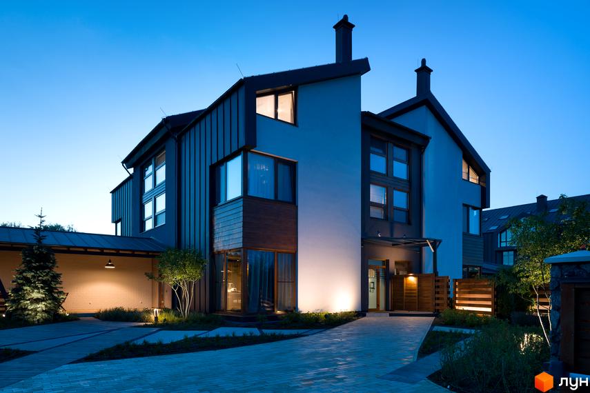 Двоповерховий будинок з панорамними вікнами та темною покрівлею. Перед будинком вимощена бруківкою доріжка, є зелені насадження та вуличне освітлення.