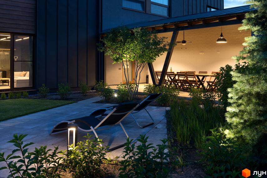 Терасований будинок з темним фасадом. На прибудинковій території розташовані шезлонги, ліхтарі та зелені насадження.