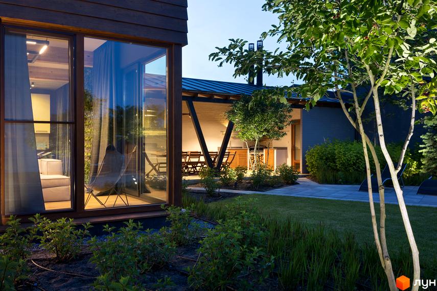 Терасований будинок з великими вікнами та дерев'яною обробкою фасаду. На прибудинковій території газон, дерева та освітлення.