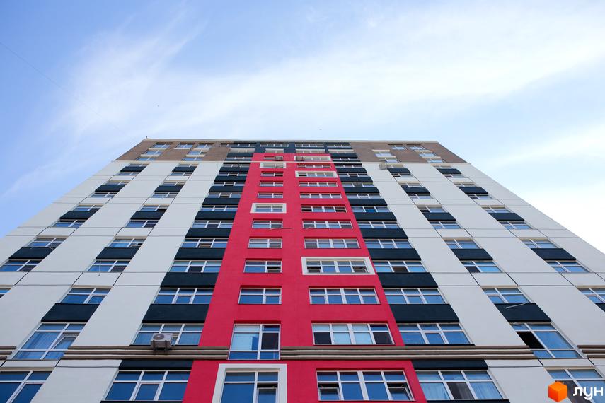 Вигляд на фасад багатоповерхового будинку Житлового комплексу Синергія-3. Будівля має червону вертикальну вставку та білі секції з численними вікнами.