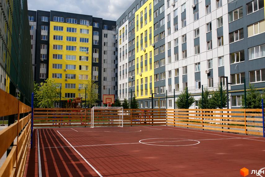 Житловий комплекс Синергія-3 з багатоповерховими будинками у жовтому, темно-сірому та світло-сірому кольорах. На території є спортивний майданчик.