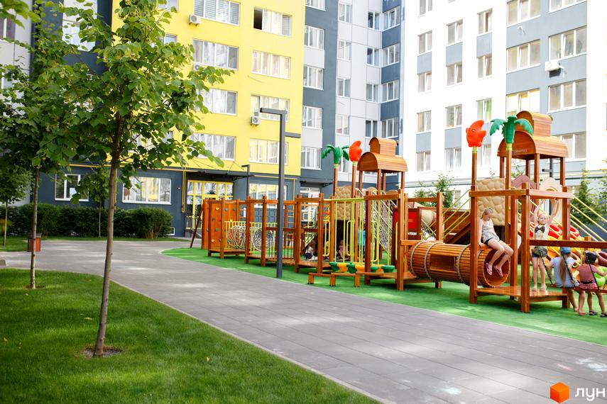Багатоповерхові будинки Житлового комплексу Синергія-3 з жовтими, сірими та білими фасадами. На передньому плані дитячий майданчик з дерев'яними елементами, зелений газон та доріжки.