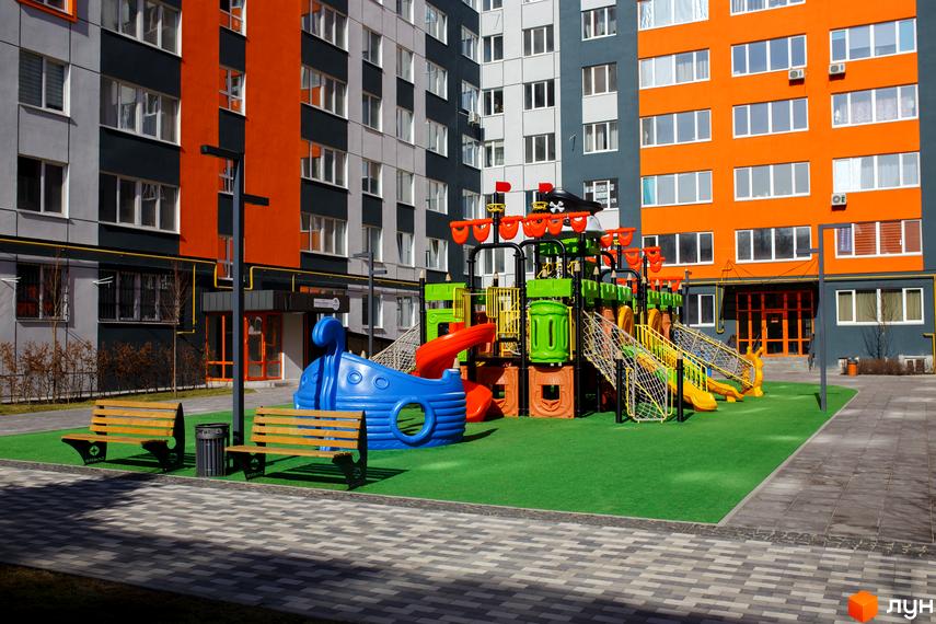 Житловий комплекс Синергія-3: багатоповерхові будинки сірого, темно-сірого та оранжевого кольорів. На подвір'ї яскравий дитячий майданчик з гірками та лавки для відпочинку.