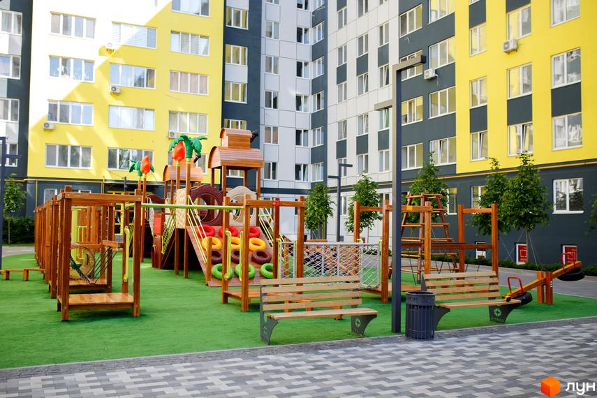 Житловий комплекс Синергія-3 з яскравими жовтими та темно-сірими фасадами. На прибудинковій території розташований великий дитячий майданчик з дерев'яними елементами та зеленим покриттям.