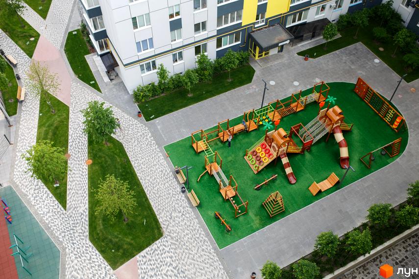 Багатоповерховий будинок Житлового комплексу Синергія-3 з сіро-жовтим фасадом. На прибудинковій території розташований великий дитячий майданчик з зеленим покриттям та ігровими елементами.