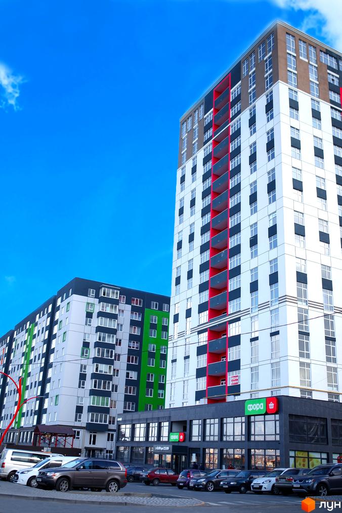 Житловий комплекс Синергія-3: високий будинок з білими фасадами, червоними балконами та темними вікнами. На першому поверсі магазин, поруч парковка з авто.