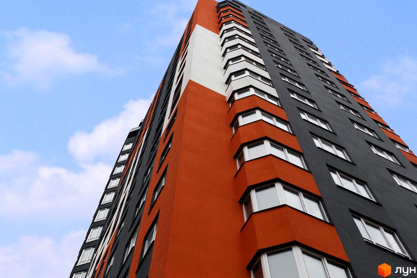 Фасад будинку ЖК Синергія-2+ з помаранчевими та сірими елементами, білою вставкою та численними вікнами на тлі блакитного неба з хмарами.