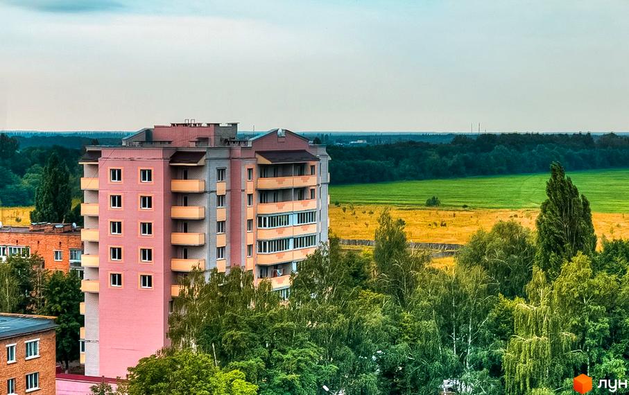 Новобудова на вулиці Вокзальна, 11а. Багатоповерховий будинок з рожевим фасадом та балконами на тлі зеленого поля та лісу.