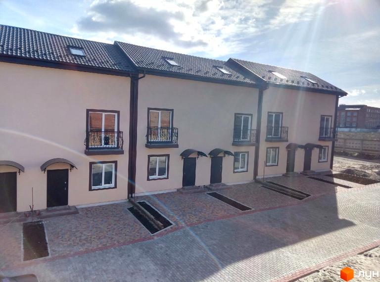 Зовнішній вигляд таунхаусів Cherry Town з сірим дахом та світлими стінами, з балконами та вікнами, на тлі хмарного неба. Поруч вимощена бруківкою прибудинкова територія з паркувальними місцями.