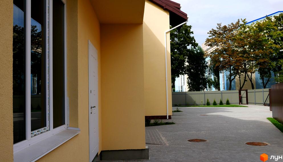 Таунхаус Нова Еллада має світло-жовті стіни та білі вікна з дверима. Прибудинкова територія викладена сірою бруківкою, огороджена парканом, з деревами та кущами.