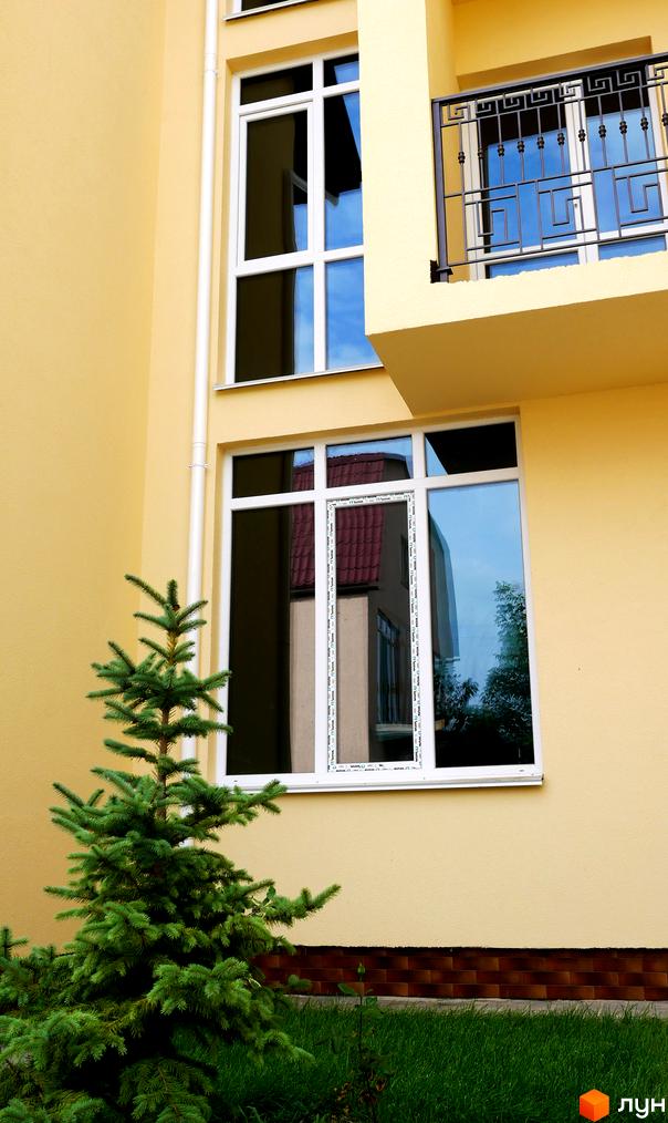 Жовтий фасад Таунхаус Нова Еллада з білими вікнами та балконом. Перед будинком росте зелена ялинка на газоні.