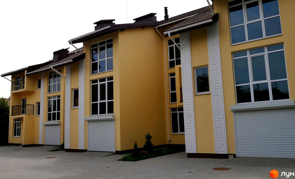 Таунхаус Нова Еллада: жовті будинки з білими вікнами, ролетними воротами та вертикальним грецьким орнаментом. Перед ними бруківка та зелені насадження.