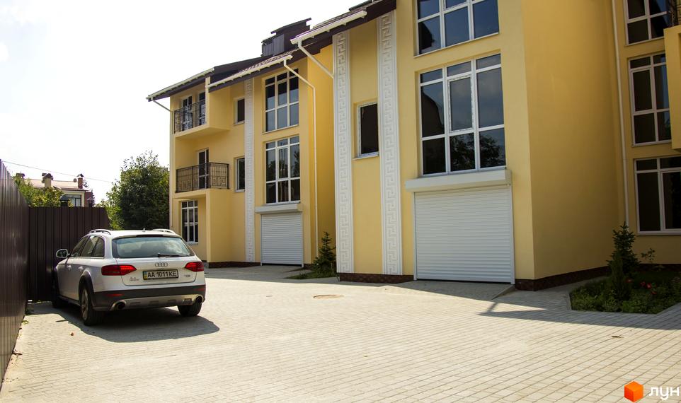 Таунхаус Нова Еллада з жовтими фасадами, великими вікнами та білими ролетними воротами гаражів. Прибудинкова територія вимощена світлою бруківкою.