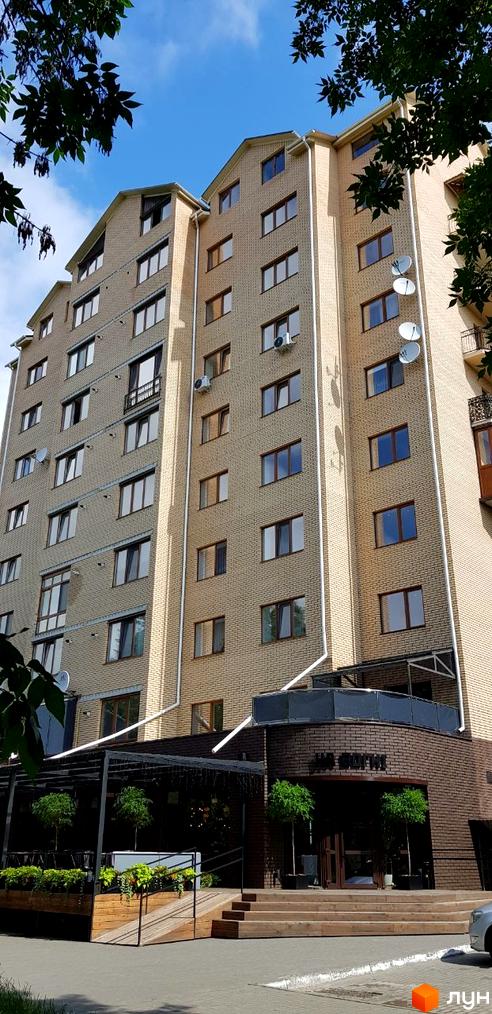 Зовнішній вигляд новобудови просп. Соборності, 16в. Фасад будинку оздоблений світлою цеглою, багато вікон та балконів. Перед будинком розташована прибудинкова територія з деревами та місцями для відпочинку.