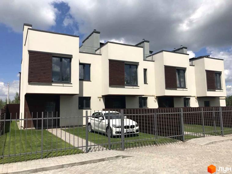 Таунхауси Monaco: ряд будинків зі світлими фасадами, темними вікнами та дерев'яними елементами. Перед будинками зелені газони та металевий паркан.