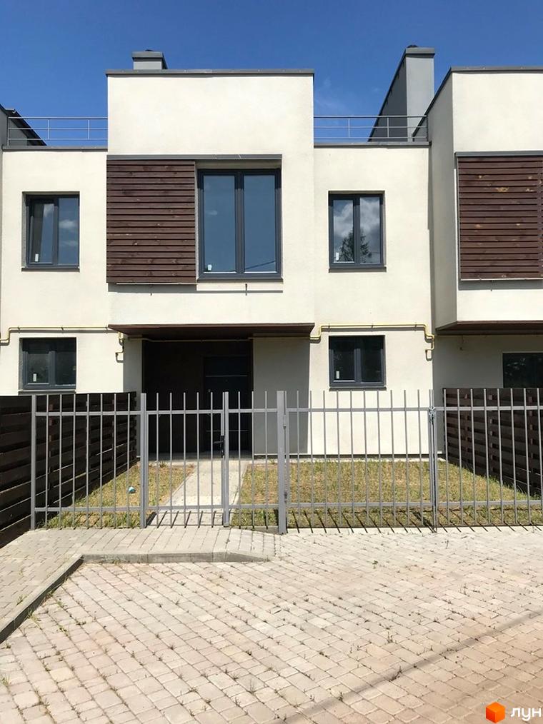 Фасад Таунхаусів Monaco з білими стінами, темними вікнами та дерев'яними панелями. На прибудинковій території сірий паркан, газон та бруківка.