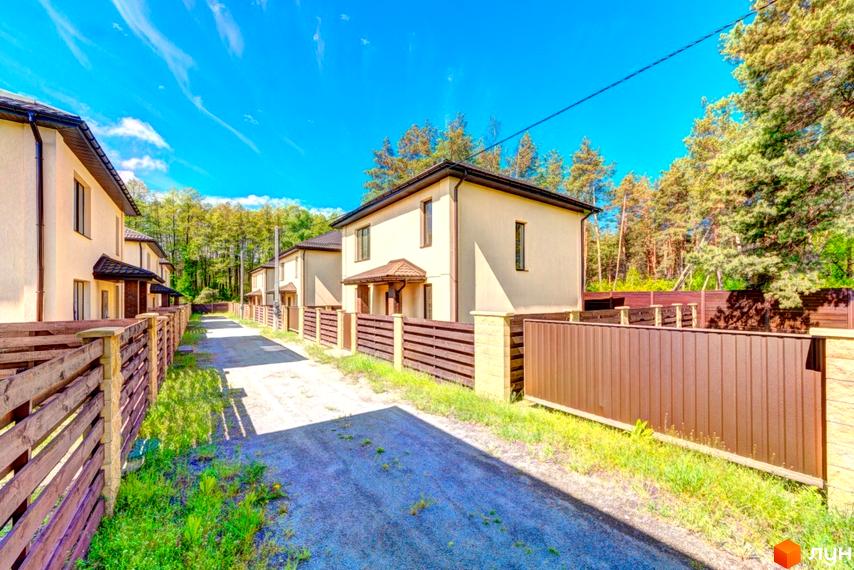 Двоповерхові будинки світлого кольору з темними дахами у Котеджному містечку Garmonia, з ґрунтовою дорогою та дерев'яними парканами.