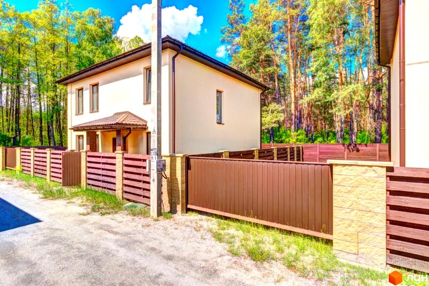 Двоповерховий будинок Котеджного містечка Garmonia світлого кольору з коричневим дахом. Територія огороджена коричневим парканом, поруч ліс.