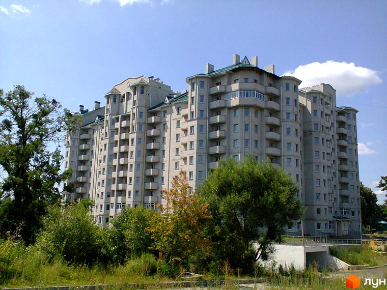 Багатоповерховий будинок сірого кольору з балконами. Поруч зелені насадження та блакитне небо.