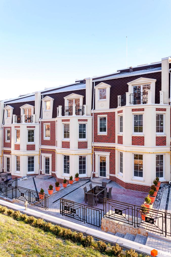 Фасад таунхаусів Royal Hill з цегляним оздобленням та великими вікнами. Прибудинкова територія з терасою, столиком, стільцями та декоративними рослинами.