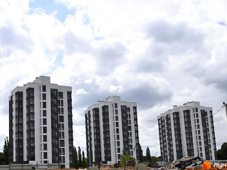 Житловий комплекс Супернова: три сучасні багатоповерхові будинки з білими та сірими фасадами, великою кількістю вікон. На передньому плані видно будівельне сміття та паркан.