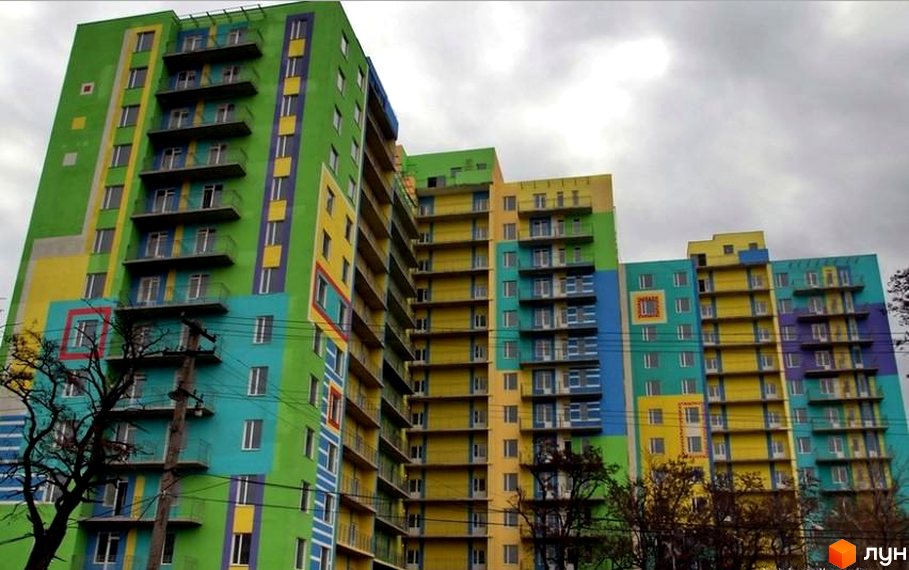 Фасад Житлового комплексу Іспанський, розфарбований у зелений, жовтий, блакитний та фіолетовий кольори, з численними вікнами та балконами. На передньому плані видно дерева.