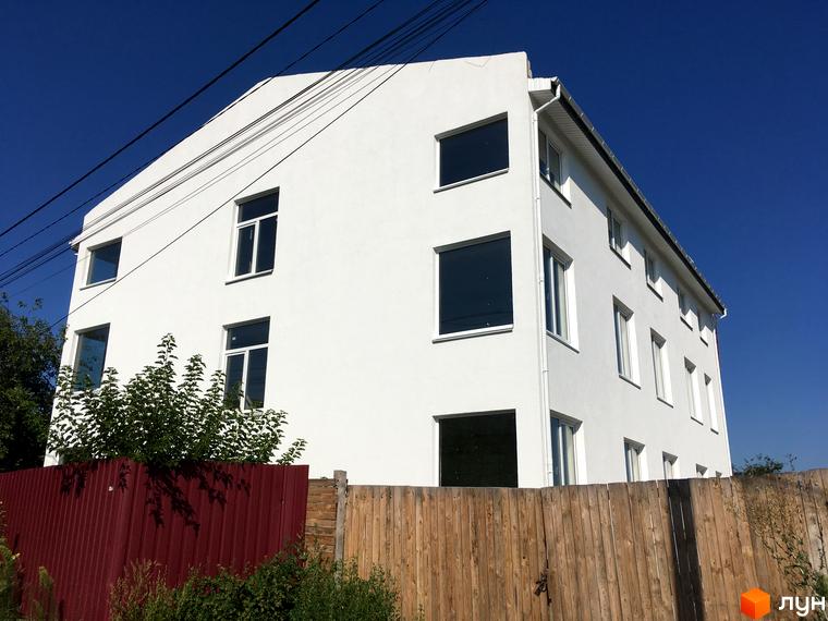 Білий багатоповерховий будинок Клубний будинок „Женева“ з численними вікнами. Перед ним розташовані червоний та дерев'яний паркани, а також зелені кущі.