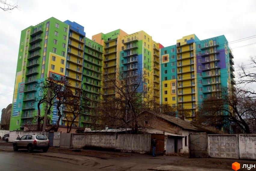 Фасад Житлового комплексу Іспанський з різнокольоровими секціями та балконами. Поруч ґрунтова дорога, дерева та паркан.