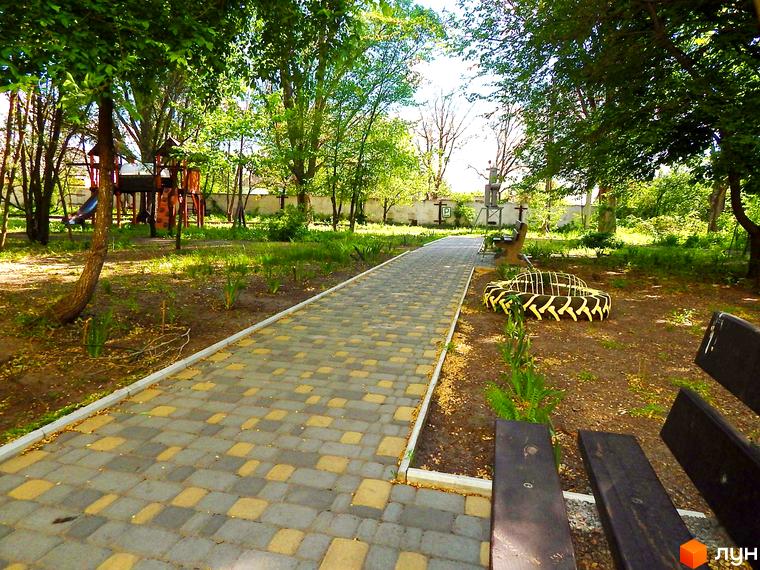 Житловий комплекс Усадьба: доглянута прибудинкова територія з пішохідною доріжкою та дитячим майданчиком. Зелені насадження та лавки для відпочинку створюють затишну атмосферу.