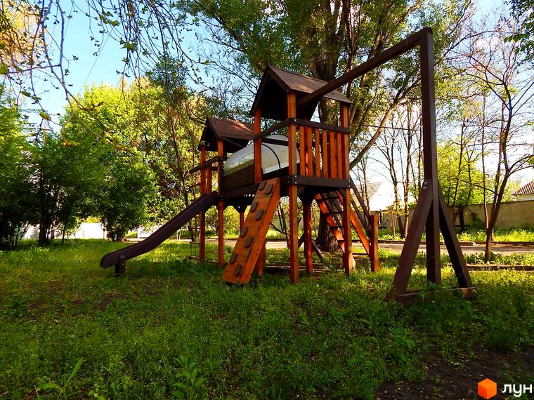 Дитячий майданчик на території ЖК Усадьба. Зелена територія з деревами та травою, дерев'яний ігровий комплекс з гіркою та драбиною.