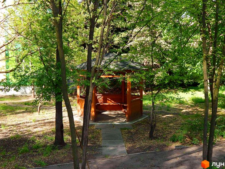 Житловий комплекс Усадьба: дерев'яна альтанка з лавками в оточенні зелені та дерев. Прибудинкова територія з доріжками та газоном.