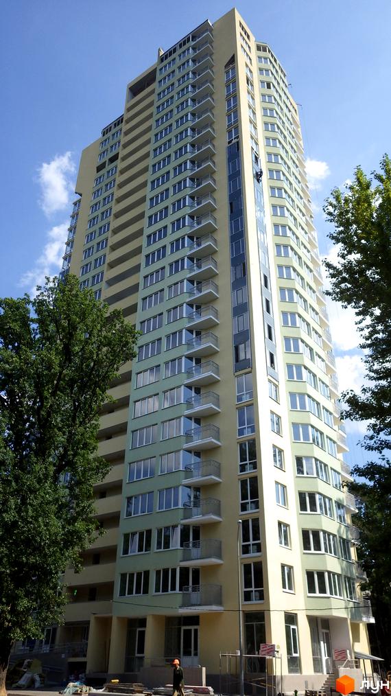 Висотна новобудова з балконами та панорамними вікнами. На передньому плані зелені дерева та блакитне небо з хмарами.