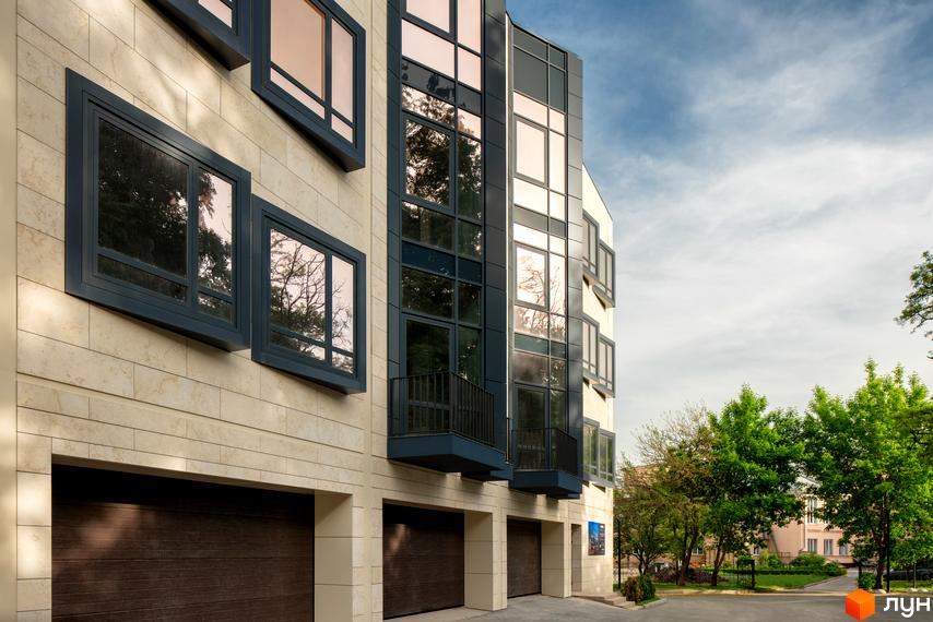 Фасад таунхаусів „Struetinsky“ зі світлих панелей, великі вікна з темними рамами та гаражі. Поруч зелені дерева.