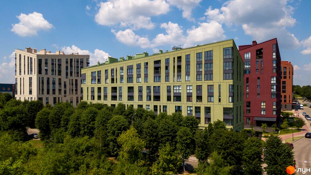 Житловий комплекс Spectrum: три багатоповерхові будинки з фасадами різних кольорів (бежевий, зелений, червоний) на тлі блакитного неба з хмарами. Навколо зелені дерева, видно дорогу з автомобілями.