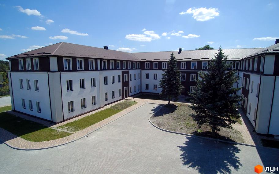 Зображення фасаду будівлі Житловий комплекс Княжий з білими стінами та коричневим дахом, з озелененням на прибудинковій території.