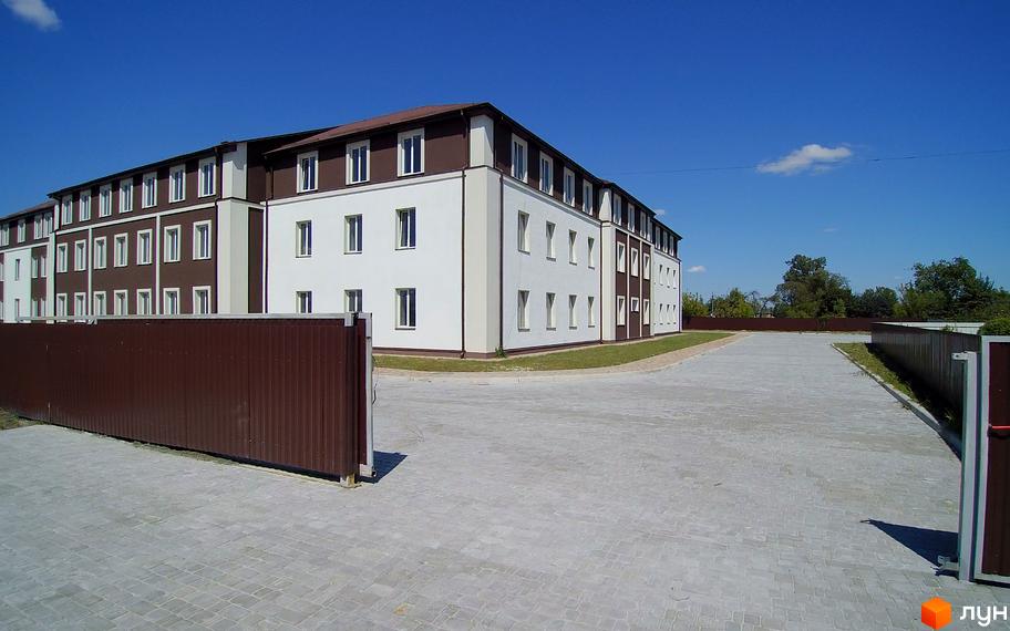 Зображення триповерхового будинку з коричневим дахом та білими стінами. На передньому плані вимощена тротуарною плиткою територія.