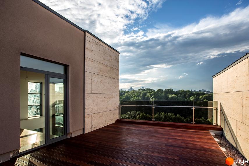 Вид на терасу житлового будинку. На терасі дерев'яна підлога, скляне огородження та вхідні двері. Поруч зелені насадження та хмарне небо.