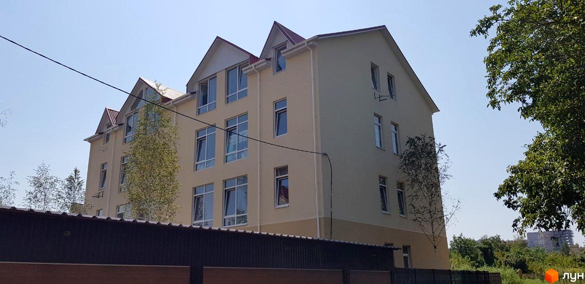 Клубний будинок „Ліон“ – фасади світло-бежевого кольору з червоним дахом, багато вікон, на тлі блакитного неба та зелені.