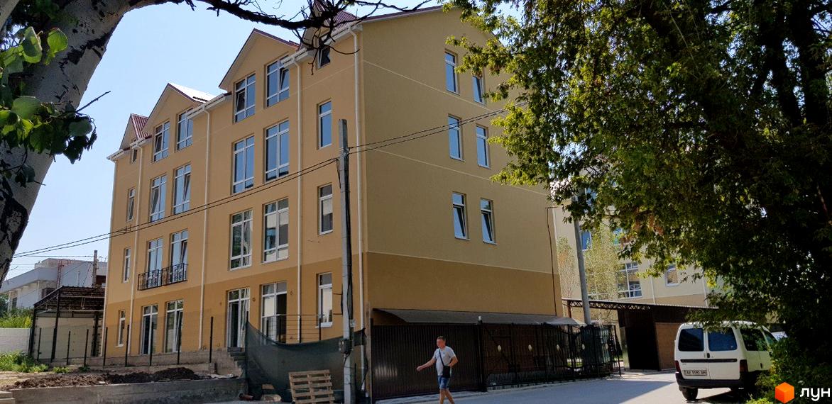 Клубний будинок Ліон – це чотириповерхова новобудова світло-бежевого кольору з великими вікнами. Територія навколо будинку огороджена парканом, поруч є парковка для автомобілів.