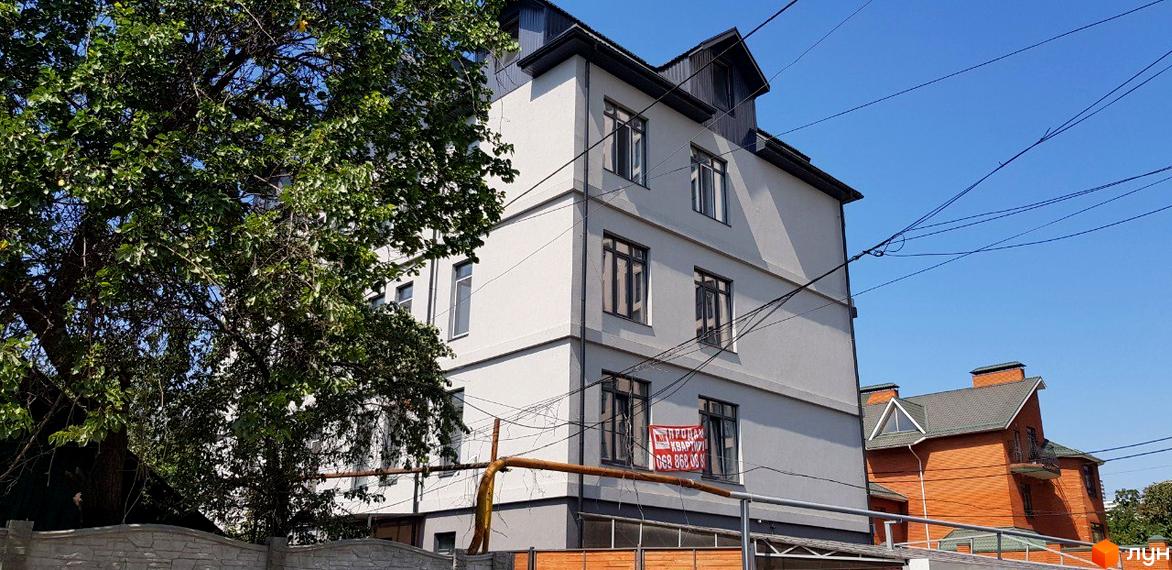 Клубний будинок „Женева“ з сірим фасадом та темним дахом, розташований біля зеленого дерева. Поруч цегляний будинок.