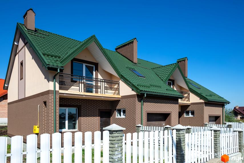 Котеджне містечко Панич. Зовнішній вигляд двоповерхових будинків з зеленим дахом, цегляними стінами та білим парканом. Навколо будівель зелений газон та блакитне небо.