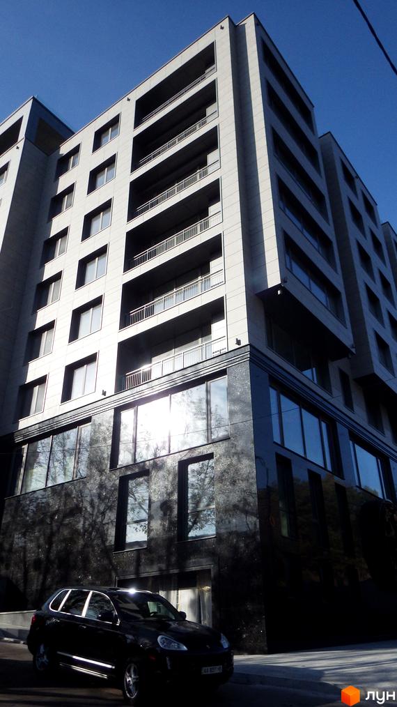 Висотна будівля житлового комплексу з великими вікнами та балконами. Перед будинком стоїть чорний автомобіль.