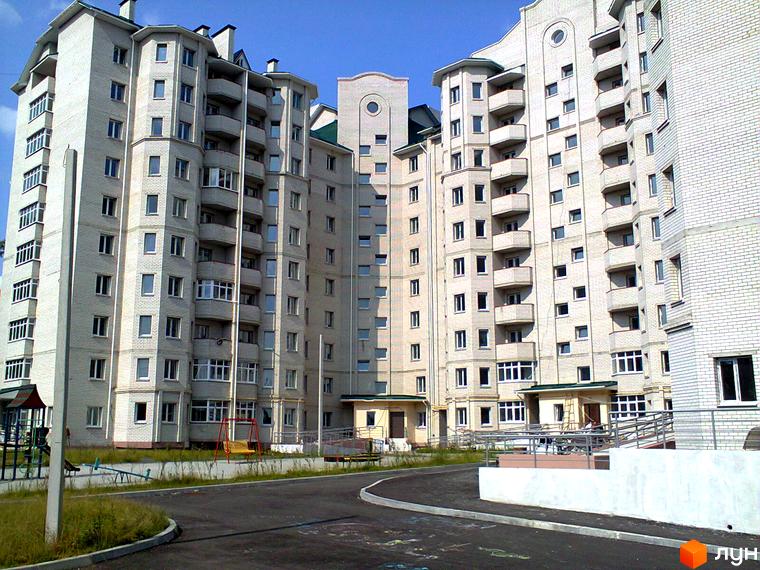 Багатоповерховий будинок з білої цегли з балконами та вікнами. На території комплексу є дитячий майданчик та дорога.