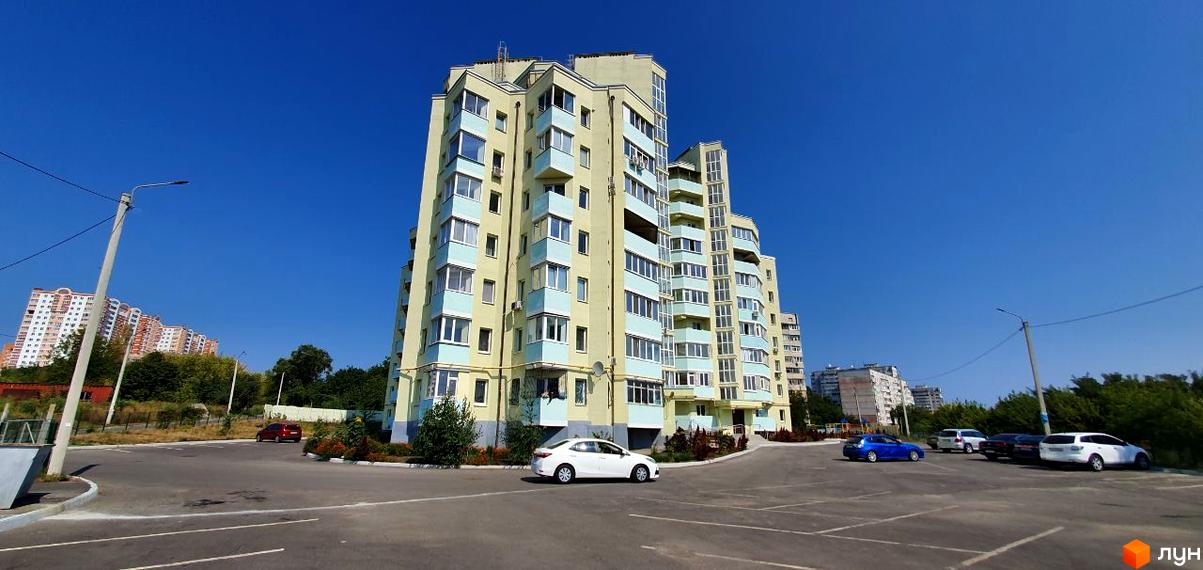 Зовнішній вигляд світло-жовтого житлового комплексу з блакитними елементами на фасаді. Поруч розташований паркінг з автомобілями та зелені насадження.