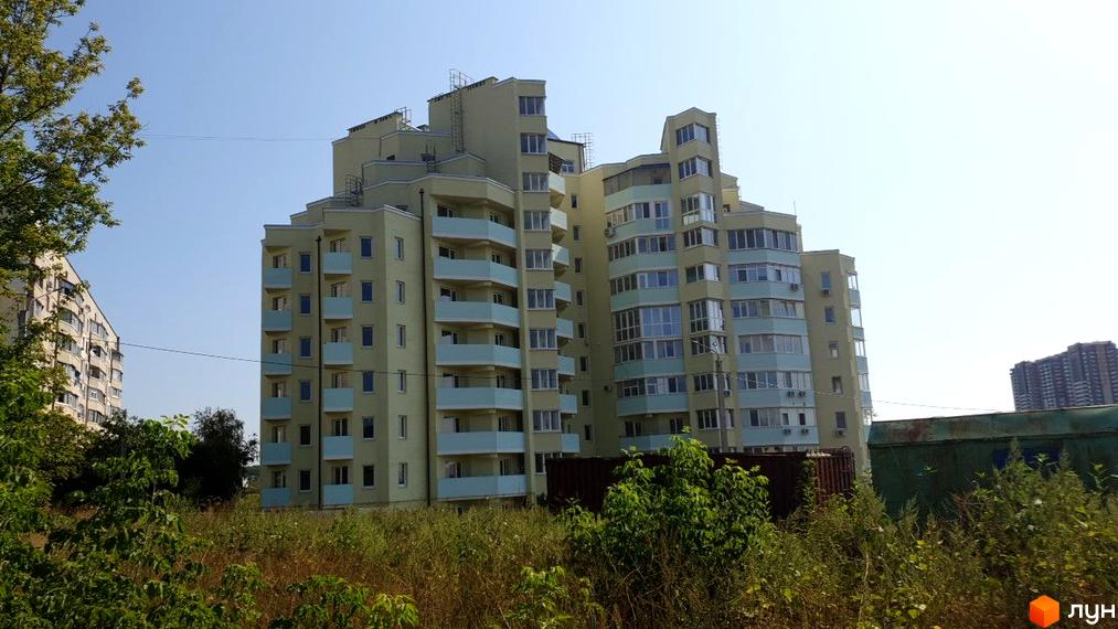 Зовнішній вигляд світло-жовтого багатоквартирного будинку з блакитними балконами. Навколо будинку зелені насадження та ділянка з травою.