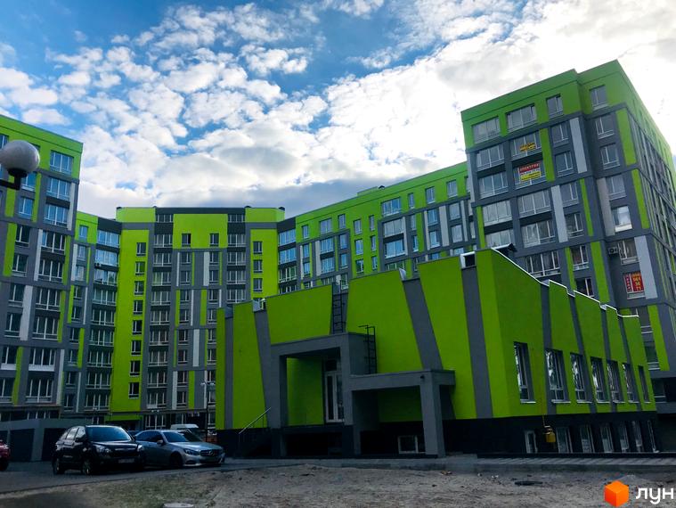 Житловий комплекс Lemongrass - яскрава новобудова з зеленими та сірими фасадами. Навколо комплексу розташовані автомобілі та ділянка з піском.