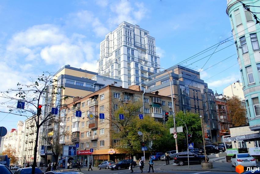 Житловий комплекс Саксаганський: багатоповерхова будівля світлих відтінків з балконами. Оточена міською забудовою, деревами та вулицею з транспортом.