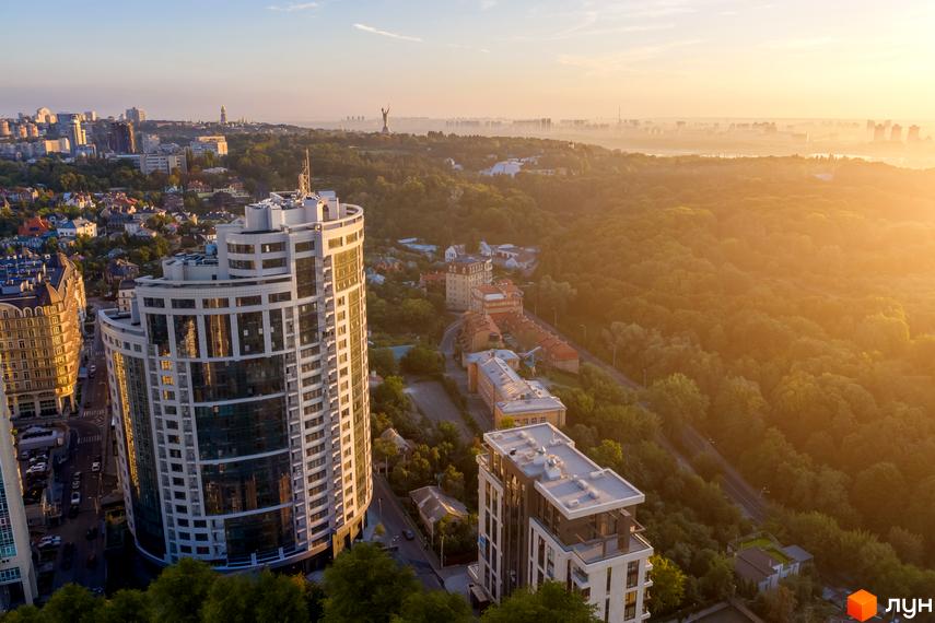 Житловий комплекс Busov Hill з білими фасадами та скляними елементами, що розташований серед зелені та міської забудови на фоні світанкового неба.